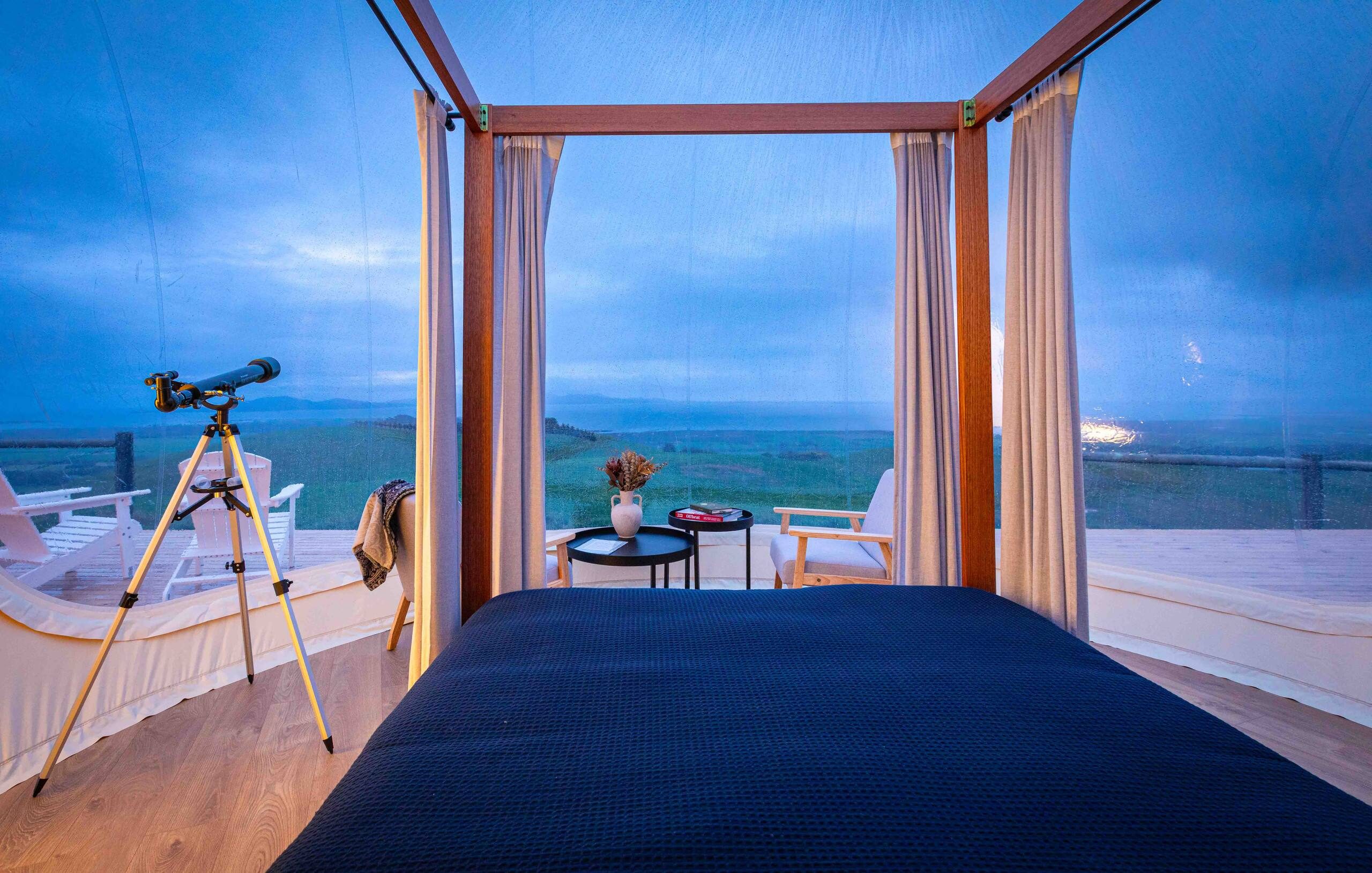 A queen bed inside the bubble with a telescope facing the panoramic view at dusk